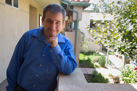 Photo of Chad Brunot standing on the balcony in front of his own home. He has a happy grin.