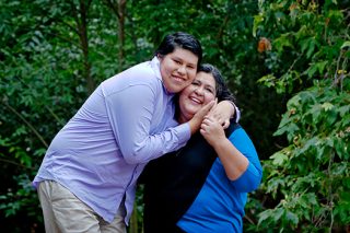 A portrait photo of a boy standing next to his mother holding her face close to his in a park. Both are smiling.