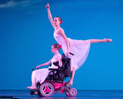 Photo of Jessica Clay in her motorized wheelchair. Jessica is wearing the flowy custume of the dancer who does an arabesque from the back of the chair.