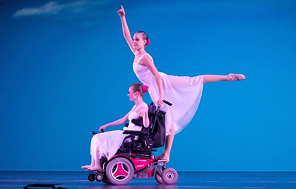 Photo of Jessica Clay in her motorized wheelchair. Jessica is wearing the flowy custume of the dancer who does an arabesque from the back of the chair.