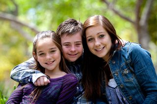 A boy with down syndrome smiles in between his two sisters while he has his arms around both of them.