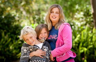 A photo of an older sister holding two smiling siblings in the park.