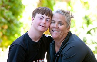 A portrait photo of a woman with her arm around a child with a developmental disability. They are both smiling.