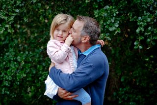 A portrait photo of a father holding his young daughter in his arms while kissing her cheek.