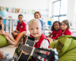 A down syndrome boy with school kids and teacher sitting on the floor in class, playing guitar.