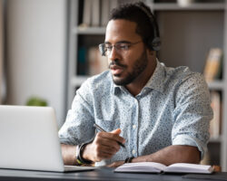 Focused, black service coordinator wears headphones as he works on his laptop in his office