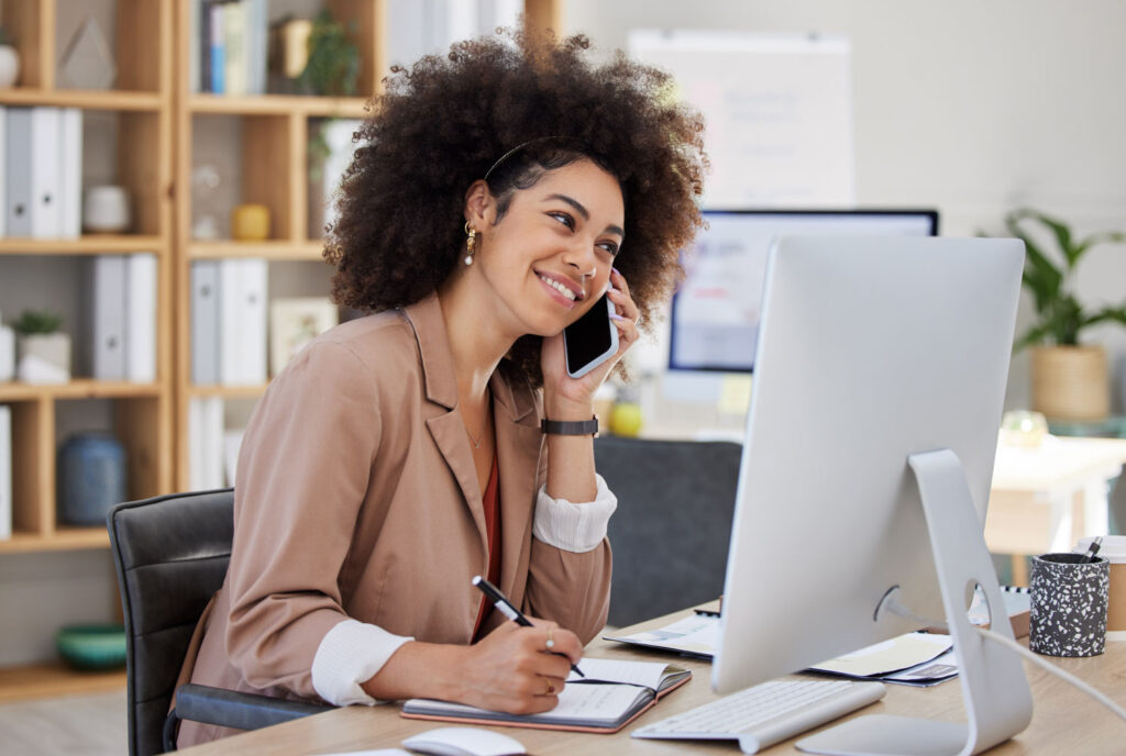 A service provider biracial person in conversation on her phone, smiles while she looks at her computer.