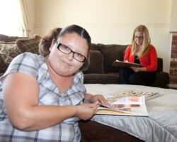 Audit of a group home with a person served in the foreground reading a book and the service provider in the background taking notes