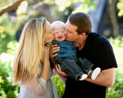 Parents hold their infant with Down Syndrome between them, each kissing a cheek