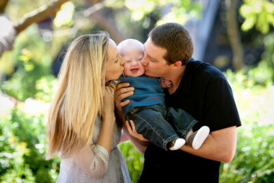 Parents hold their infant with Down Syndrome between them, each kissing a cheek