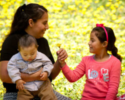 Hispanic mom holds a young son with down syndrome while her daughter hands her a yellow flower