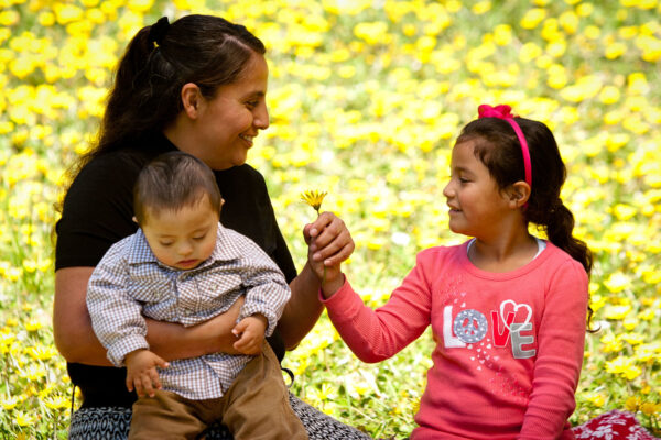 Hispanic mom holds a young son with down syndrome while her daughter hands her a yellow flower