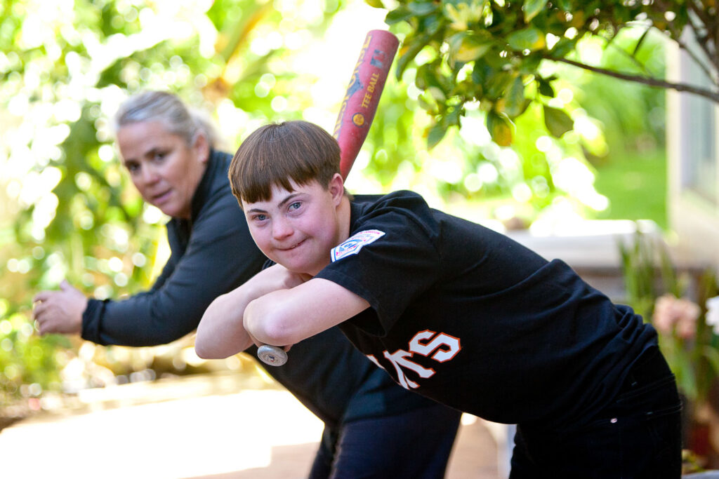 Young man with Down Syndrome practices holding a baseball bat with his care-giver