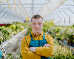 Young employee with Down syndrome working in garden centre, looking at camera with arms crossed.