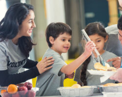 Elementary age Hispanic boy is serving meal to senior man in charity soup kitchen at food bank. His mother, father, and sister are with him. Family is volunteering for charity together. They are wearing VOLUNTEER t-shirts and serving healthy food.