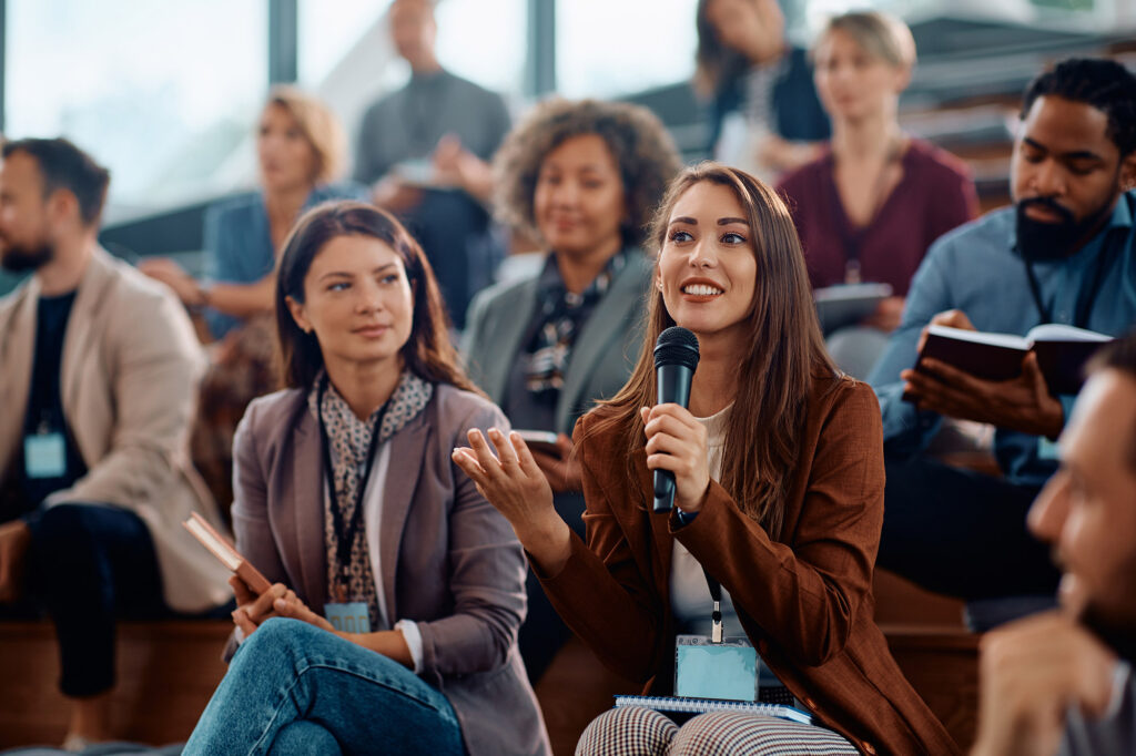 Woman holds a microphone in a diverse audience as she asks a question.