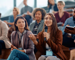 Woman holds a microphone in a diverse audience as she asks a question.